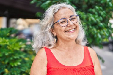 Orta yaşlı, kır saçlı, gülümseyen, kendine güvenen bir kadın parkın kenarına bakıyor.