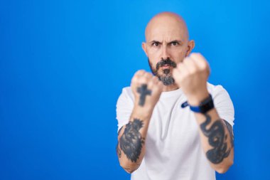 Mavi arka planda dikilen İspanyol dövmeli adam yumruk savunma hareketleriyle dövüşmeye hazır kızgın ve üzgün yüzlü, sorundan korkan. 