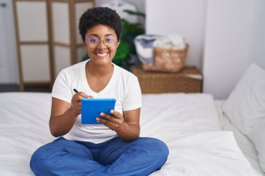 African american woman using touchpad sitting on bed at bedroom