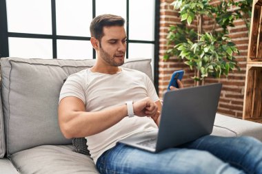 Young hispanic man using laptop and laptop looking watch at home