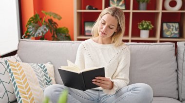 Young blonde woman reading book sitting on sofa at home