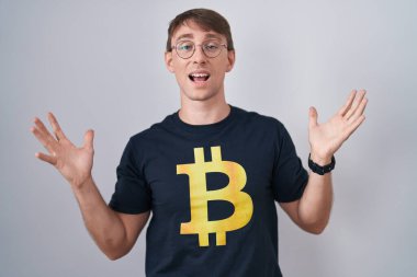 Bitcoin tişörtü giyen beyaz, sarışın bir adam çılgın kutlamalar yapıyor ve kolları açık, gözleri açık heyecanla haykırıyor. Kazanan konsept 
