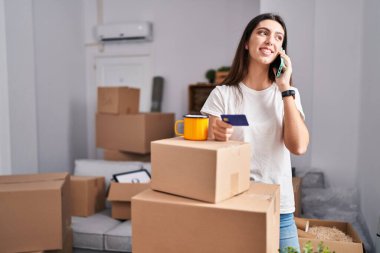 Young beautiful hispanic woman talking on smartphone holding credit card at new home