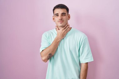 Yakışıklı İspanyol bir adam pembe arka planda duruyor. Acı veren boynuna dokunuyor. Grip, mantar ve enfeksiyon yüzünden boğazı ağrıyor. 