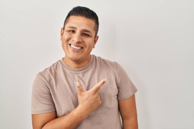 Latin kökenli genç bir adam beyaz arka planda neşeli, yüzünde mutlu ve doğal bir ifadeyle elleri ve parmaklarıyla işaret eden bir gülümsemeyle dikiliyor. 