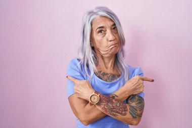 Pembe arka planda duran orta yaşlı kadın parmaklarıyla her iki tarafı işaret ediyor, farklı yönlere bakıyor. 