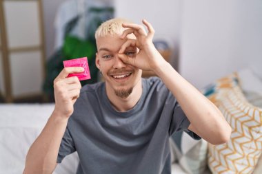 Kafkasyalı genç adam, elinde prezervatif, yatağında oturmuş gülümsüyor, mutlu mesut gülümsüyor. 