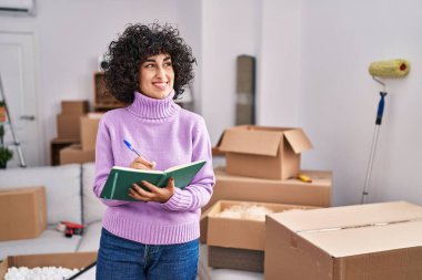 Orta Doğulu genç bir kadın yeni evinde deftere yazarak gülümsüyor.