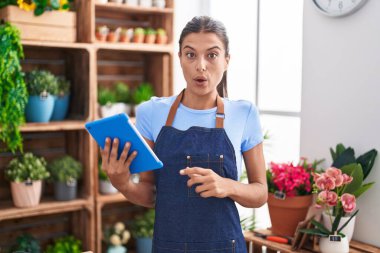 Çiçekçi dükkanında çalışan esmer genç bir kadın elinde tabletle korkmuş ve ağzı açık şaşkınlık içinde şaşkınlık içinde, inançsız bir yüz. 