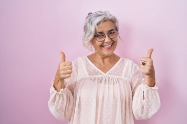 Pembe arka planda duran gri saçlı, elinde olumlu bir jest yapan mutlu ve gülümseyen orta yaşlı bir kadın. Neşeli ifade ve kazanan jest. 