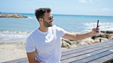 Young hispanic man smiling confident making selfie by the smartphone at seaside