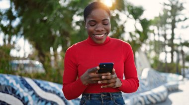 Parkta gülümseyen akıllı telefon kullanan Afrikalı Amerikalı kadın.