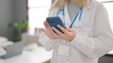 Klinikte akıllı telefon kullanan genç sarışın kadın doktor.