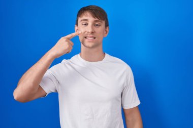 Mavi arka planda dikilen beyaz, sarışın bir adam. Parmakla yüzünü ve burnunu gösteriyor. Neşeli bir şekilde gülümsüyor. güzellik konsepti 