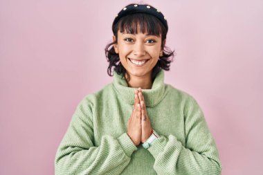 Young beautiful woman standing over pink background praying with hands together asking for forgiveness smiling confident. 