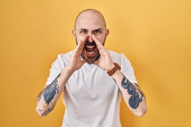 Sarı arka planda dikilen dövmeli genç İspanyol adam elleri ağzında bağırarak bağırıyor. 