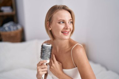 Young blonde woman combing hair sitting on bed at bedroom