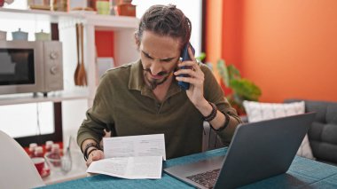 Genç İspanyol adam, yemek odasında dizüstü bilgisayar okuyarak akıllı telefondan konuşuyor.