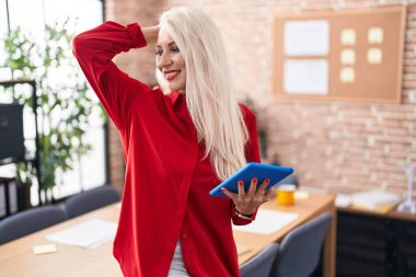 Ofiste çalışan beyaz kadın tabletle gülümsüyor saçlarına dokunuyor, el hareketi yapıyor, çekici ve modaya uygun poz veriyor. 