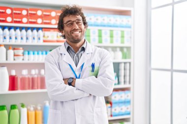 Young hispanic man pharmacist smiling confident standing with arms crossed gesture at pharmacy