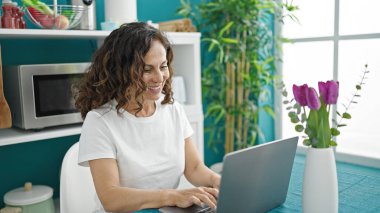 Orta yaşlı İspanyol kadın yemek odasındaki masada dizüstü bilgisayar kullanıyor.