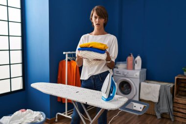 Şok olmuş yüzünü ütüledikten sonra çamaşırları katlayan esmer kadın şüpheci ve alaycı görünüyor, ağzı açık kalınca şaşırıyor. 