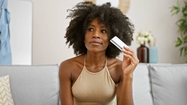 Elinde kredi kartı olan Afrikalı Amerikalı bir kadın kanepeye oturmuş, evde oturup düşünüyor.