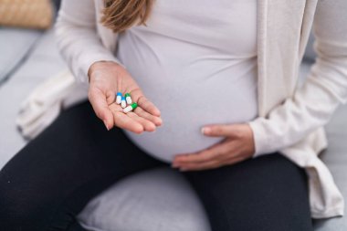 Young pregnant woman touching belly holding pills at home