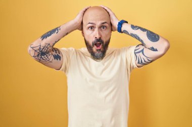 Sarı arka planda dikilen İspanyol dövmeli adam deli ve elleri başında korkmuş, ağzı açık şoktan korkmuş ve şaşırmış. 