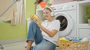 Çamaşır odasında gülümseyen akıllı telefon ve kulaklık kullanan genç sarışın kadın.