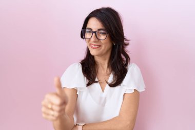 Orta yaşlı İspanyol kadın, günlük beyaz tişört ve gözlük takıyor. Gururlu görünüyor, gülümsüyor, baş parmak işareti yapıyor. 