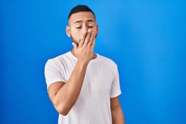 Mavi arka planda dikilen İspanyol genç adam ağzıyla ağzını kapatmaktan sıkılmış. Huzursuz ve uykulu.. 