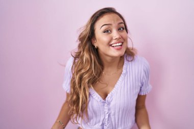 Young hispanic woman standing over pink background smiling cheerful with open arms as friendly welcome, positive and confident greetings 