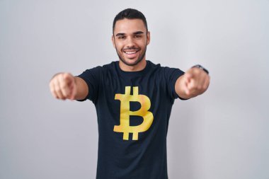 Bitcoin tişörtü giyen genç İspanyol adam seni ve kamerayı işaret ediyor, pozitif ve neşeli gülümsüyor. 