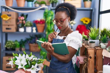 African american woman florist reading notebook with doubt expression at florist store
