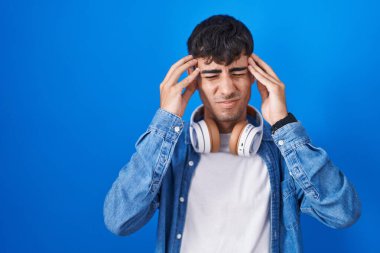 Young hispanic man standing over blue background with hand on head, headache because stress. suffering migraine. 