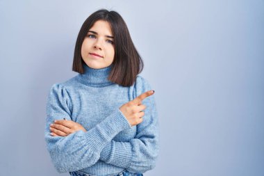 Young hispanic woman standing over blue background pointing with hand finger to the side showing advertisement, serious and calm face 