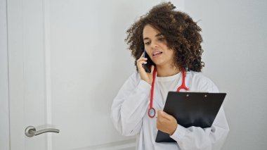 Genç, güzel İspanyol kadın doktor. Klinikte elinde not panosuyla akıllı telefondan konuşuyor.