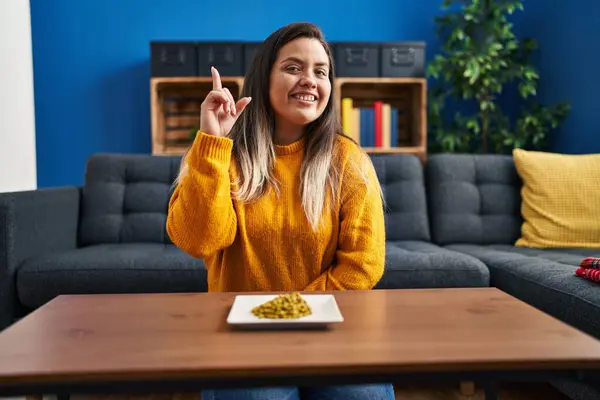 Young hispanic woman eating green peas smiling with an idea or question pointing finger up with happy face, number one