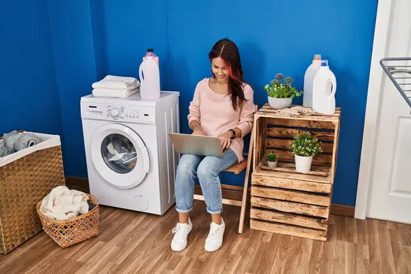 Young caucasian woman using laptop waiting for washing machine at laundry room