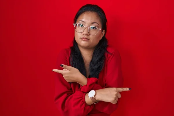 アジアの若い女性は指で両側を指している赤い背景の上に立って 異なる方向 — ストック写真