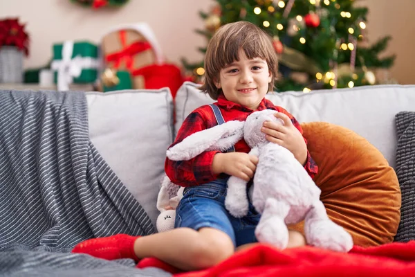 Adorable toddler hugging rabbit doll sitting on sofa by christmas tree at home