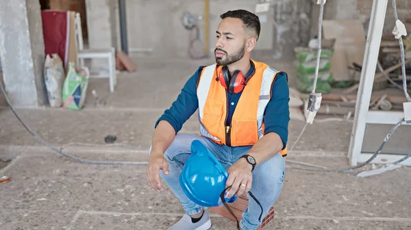 Young hispanic man builder sitting on the floor resting at construction site
