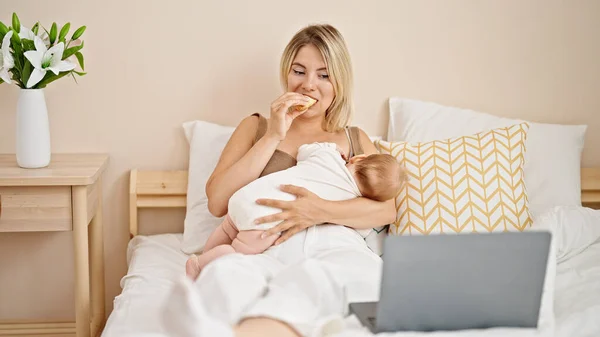 ベッドルームでクロワッサンを食べるラップトップで映画を見ている母と娘の授乳赤ちゃん — ストック写真