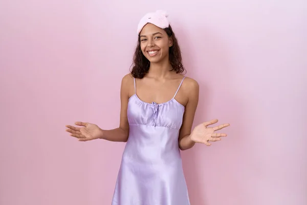 Young hispanic woman wearing sleep mask and nightgown smiling cheerful with open arms as friendly welcome, positive and confident greetings
