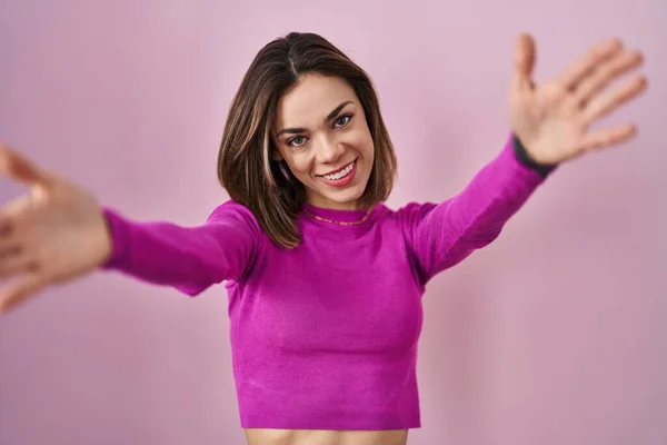 Hispanic woman standing over pink background looking at the camera smiling with open arms for hug. cheerful expression embracing happiness.