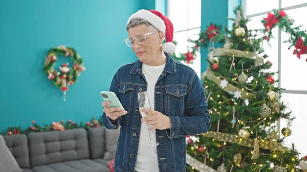 自宅でシャンパンを持っているクリスマスを祝うスマートフォンを使用する若い白人男性 — ストック写真