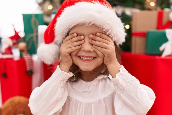 Adorable hispanic girl covering eyes with hands sitting on floor by christmas tree at home