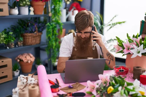 Young redhead man florist talking on smartphone using laptop at flower shop