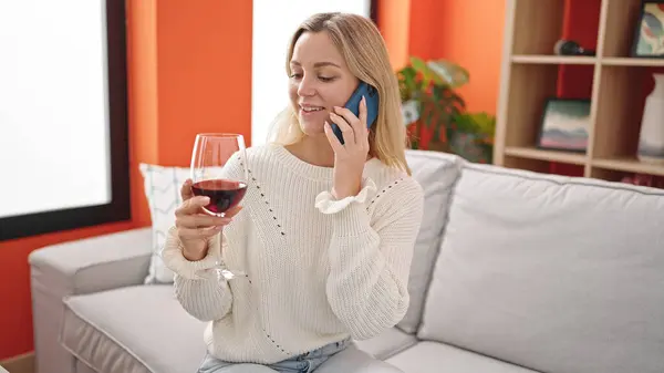 Young blonde woman talking on the smartphone and drinking wine at home